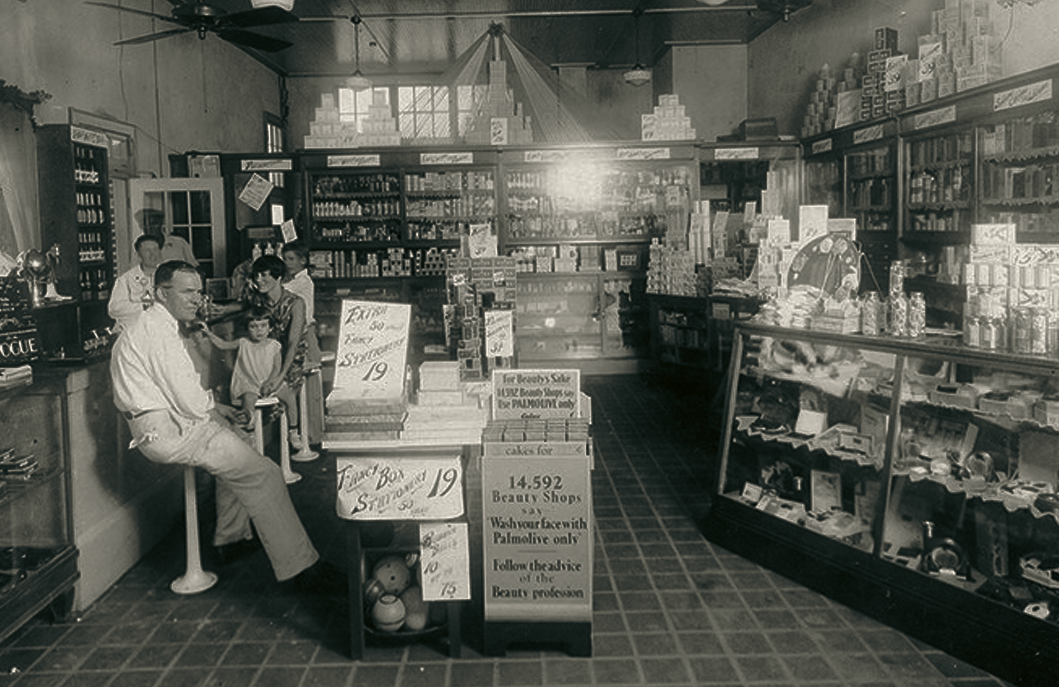 Vintage black and white photo of a classic American drugstore interior with customers, shelves of merchandise, and period advertising